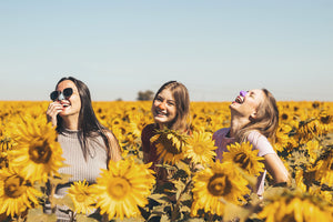 three women laughing in midst of field of sunflowers with colorful spf sunscreen on their nose